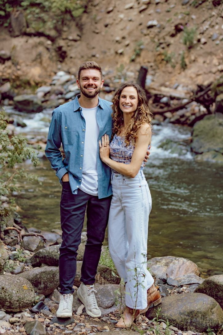 Couple posing together on rocky riverbank surrounded by nature.