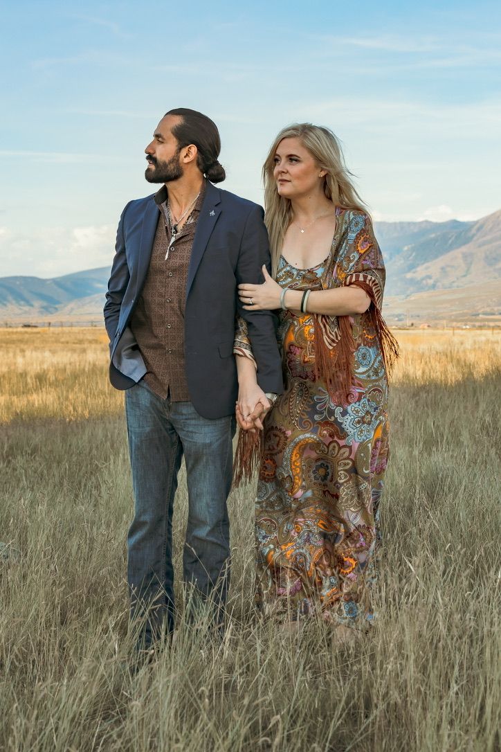 Couple walking together in field of tall grass with mountains in background.