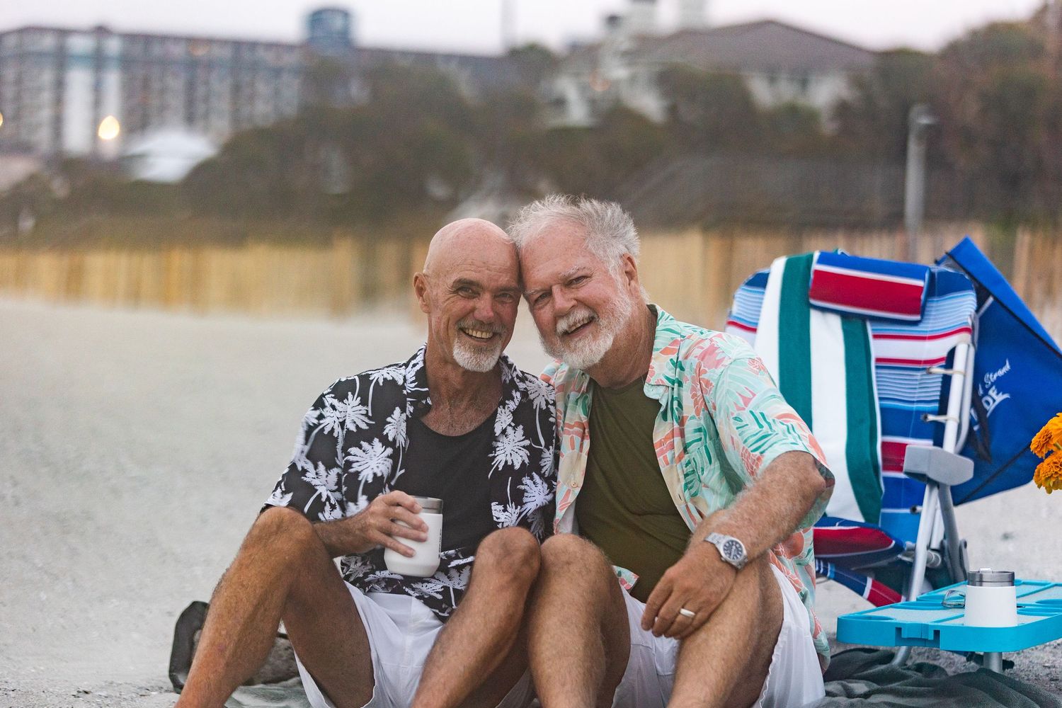 Two smiling senior men with white beards sit together on a beach, wearing colorful shirts near beach chairs.