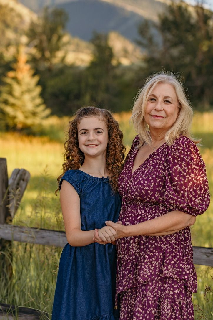 Two people in dresses pose by a wooden fence at sunset with mountains in the background.
