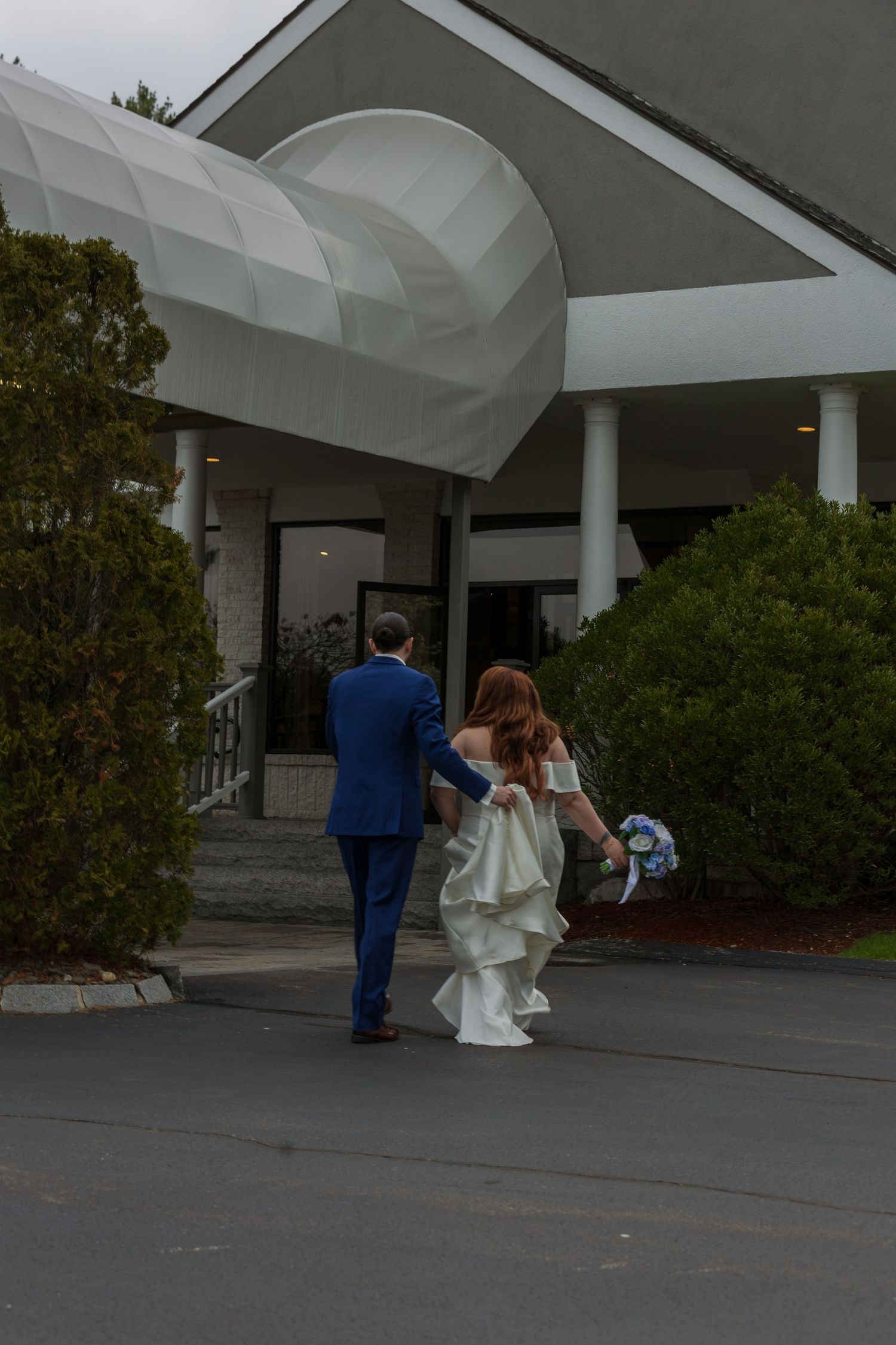 Bride and groom walking hand-in-hand toward a wedding venue entrance, bride in white gown holding bouquet, groom in blue suit.
