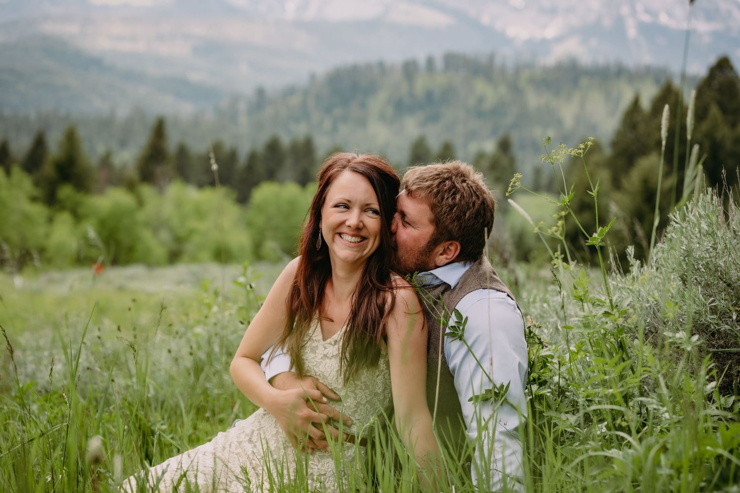 Couple shares intimate moment in wildflower meadow with mountains in background.