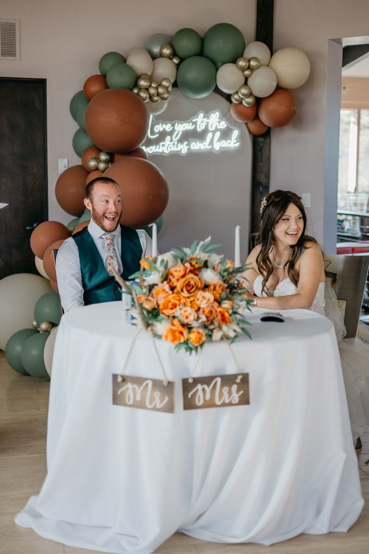 Newlyweds sit at sweetheart table with balloon arch and neon sign decor.