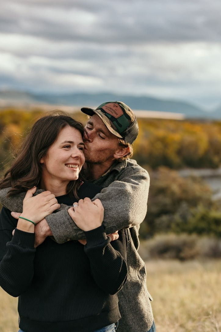 Couple embracing outdoors in autumn field with mountains and cloudy sky.