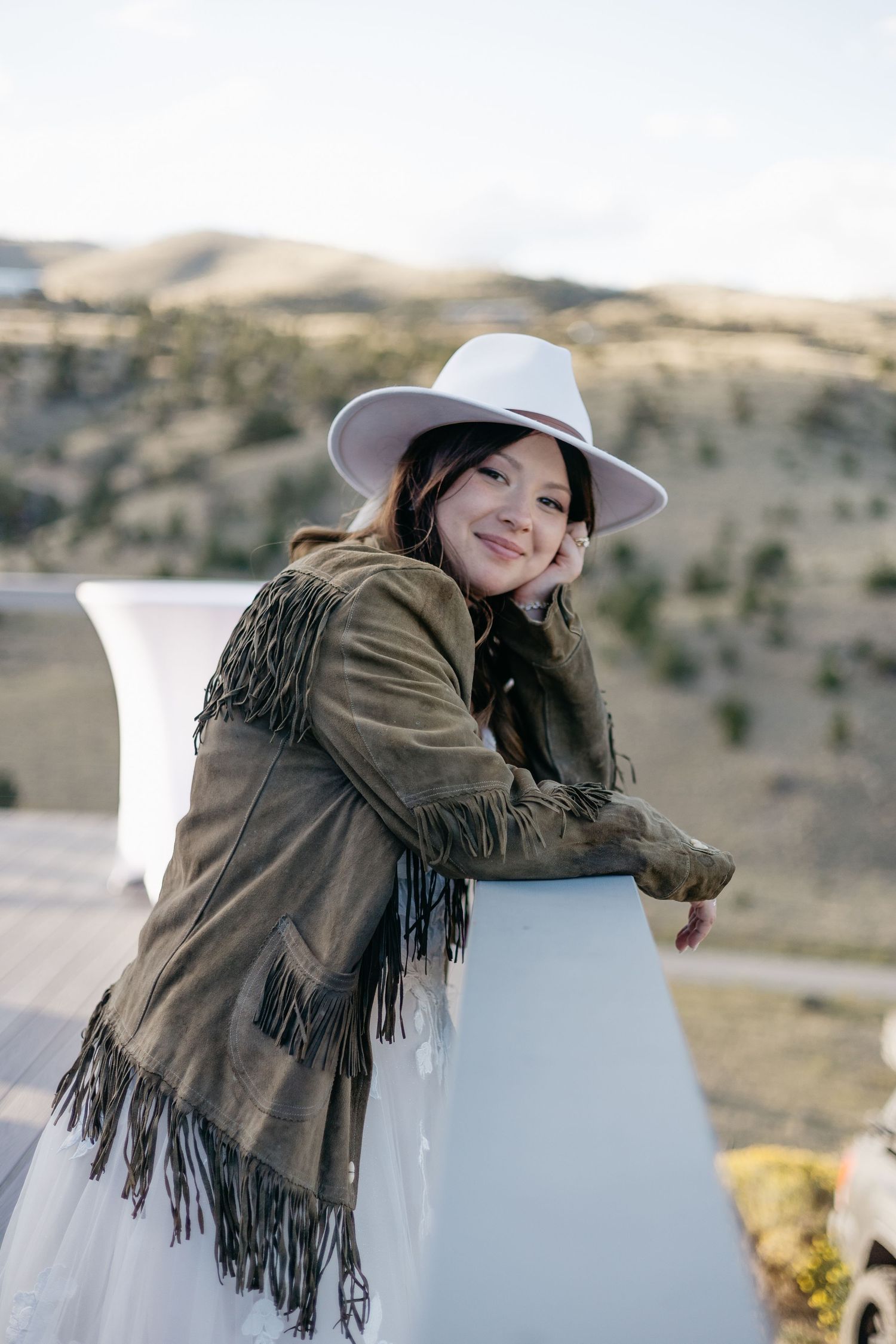 A person in a brown fringe jacket and white hat leans against a railing with mountains in the distance.