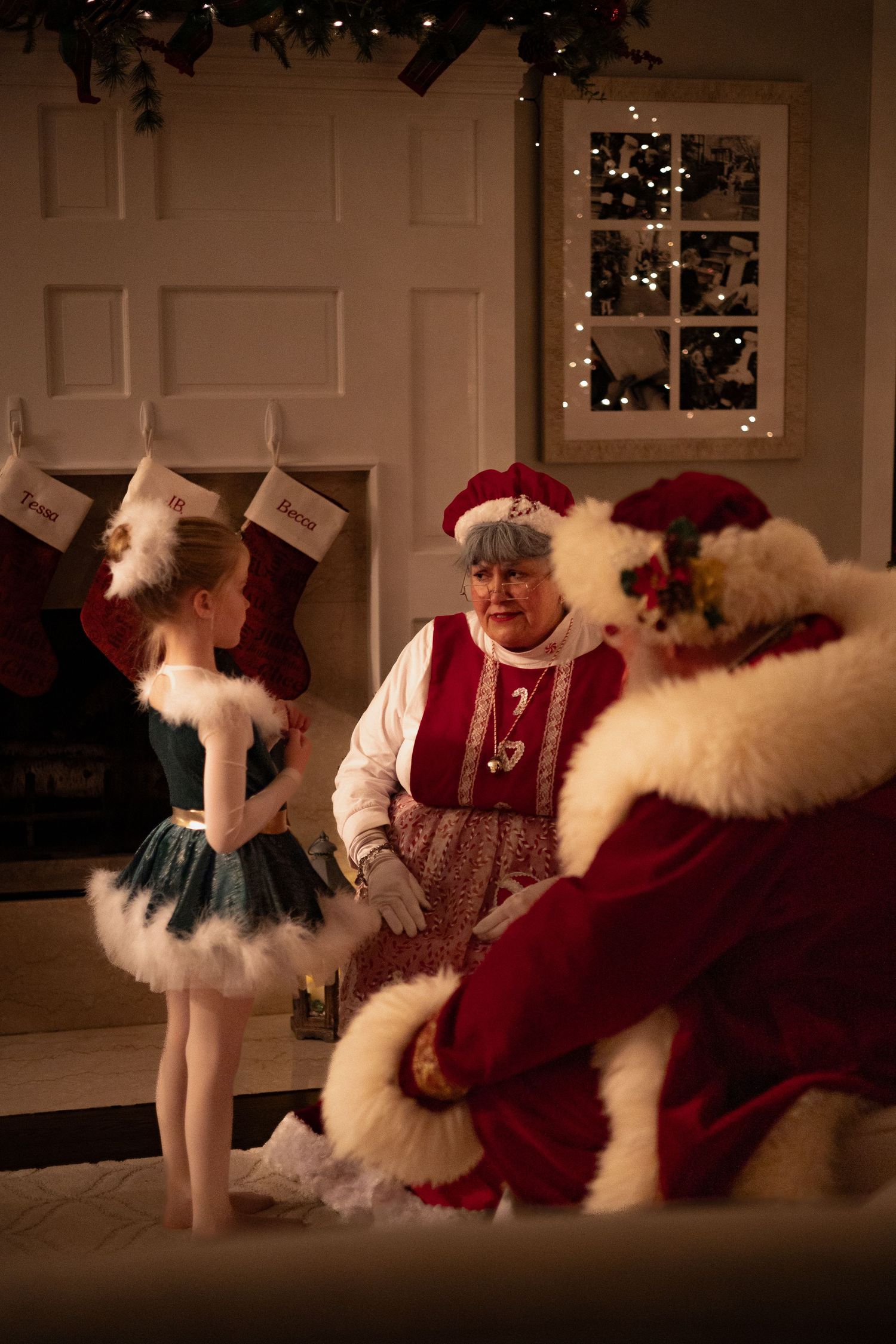 A young girl in a teal tutu meets Santa and Mrs. Claus by a decorated fireplace with stockings hung.