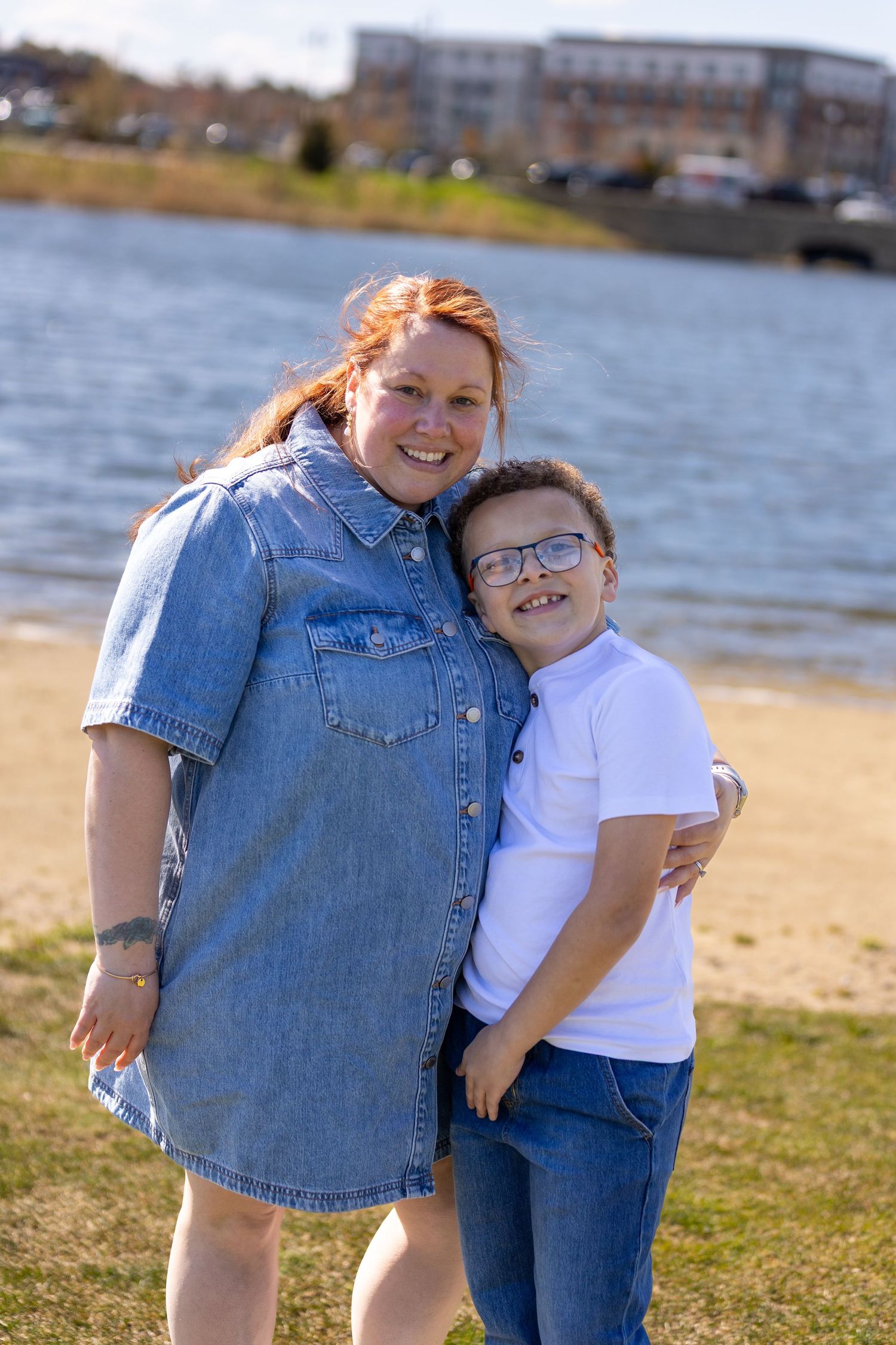 A smiling redhead woman in a denim dress hugs a young boy with glasses in a white shirt by a sunny waterfront.
