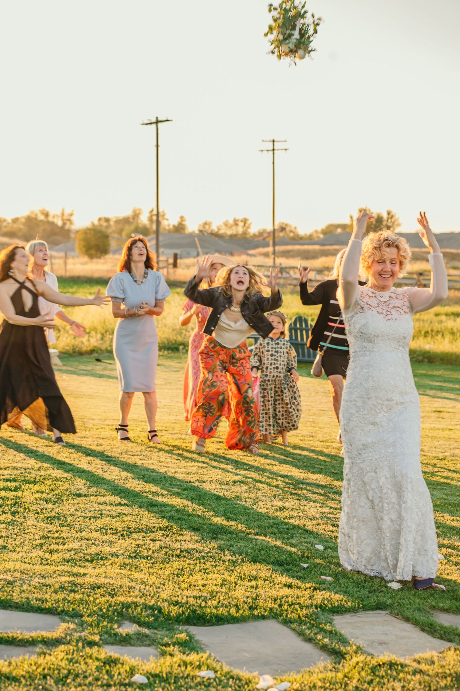 Bridal bouquet toss at sunset with excited wedding guests on a grassy lawn.