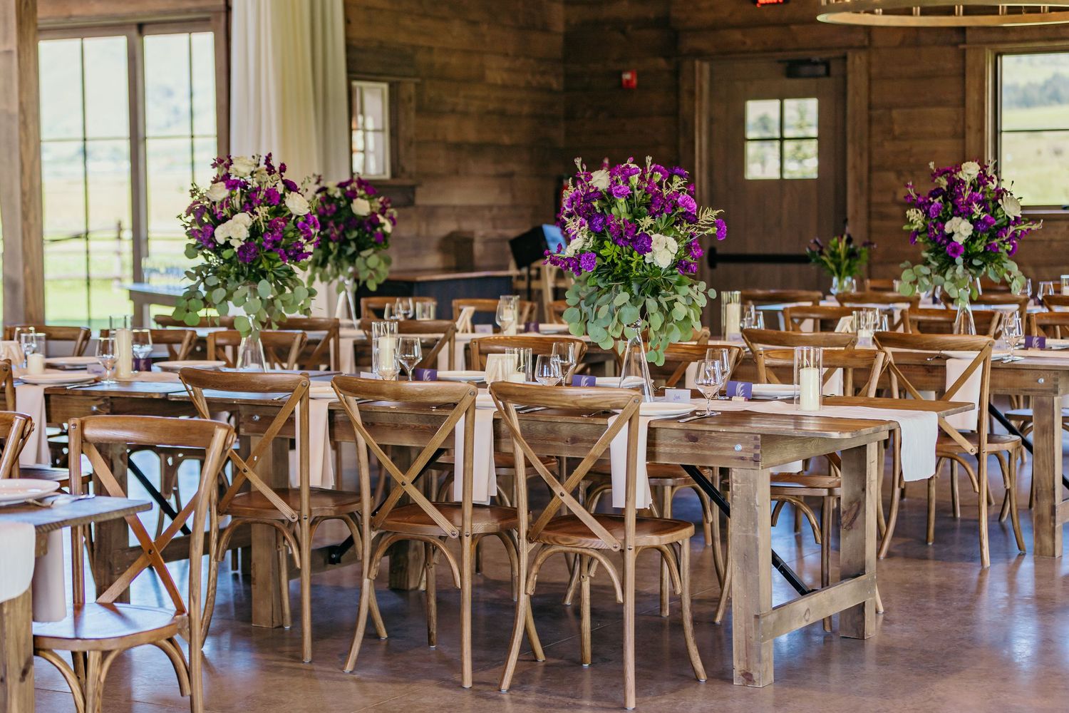 Rustic reception venue with wooden cross-back chairs and long farm tables set for dining.