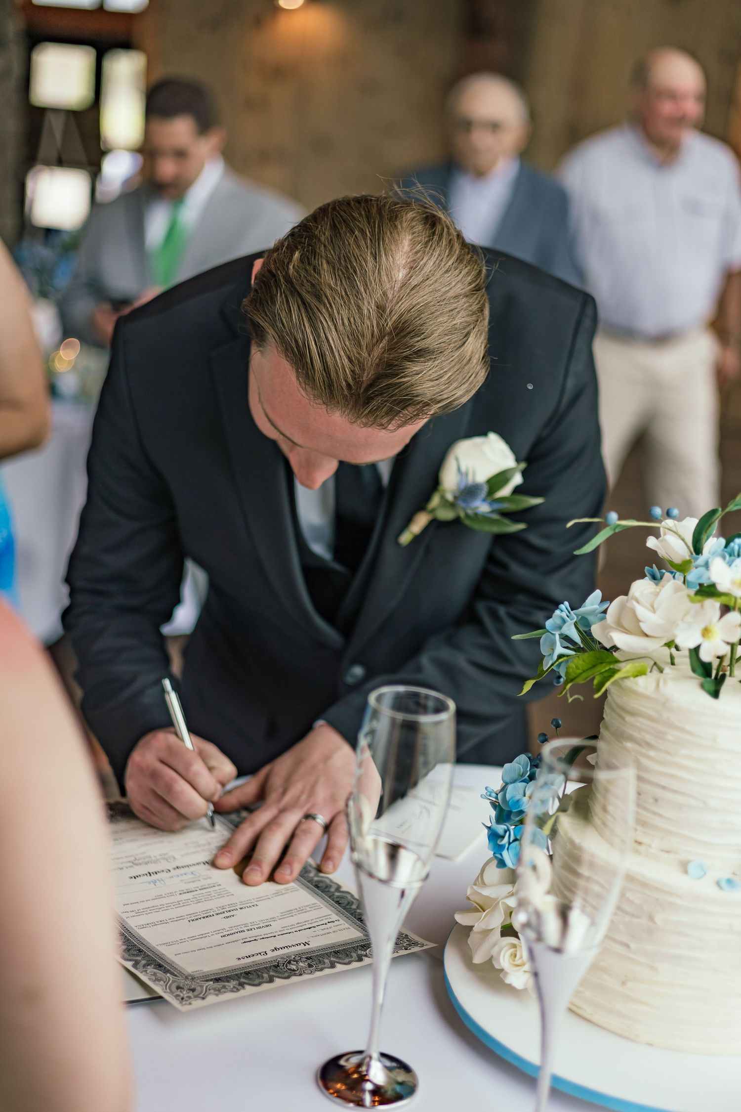 Wedding guest signs marriage certificate next to elegant white tiered cake with floral decorations.