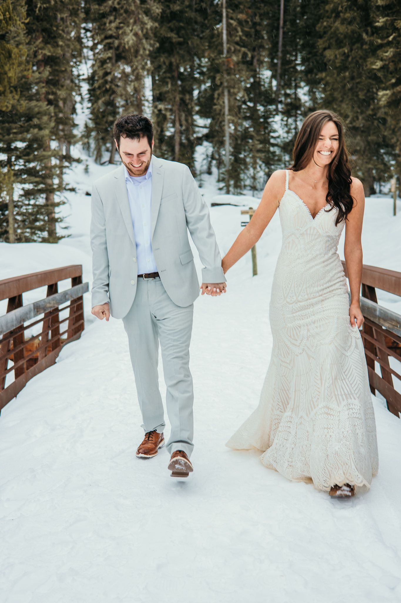 A couple walks hand in hand across a snow-covered bridge in wedding attire.