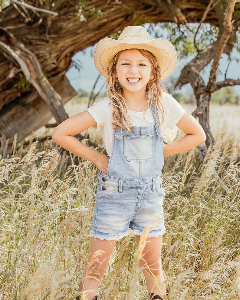 A country-style portrait in overalls and cowboy hat amid tall grass and trees.