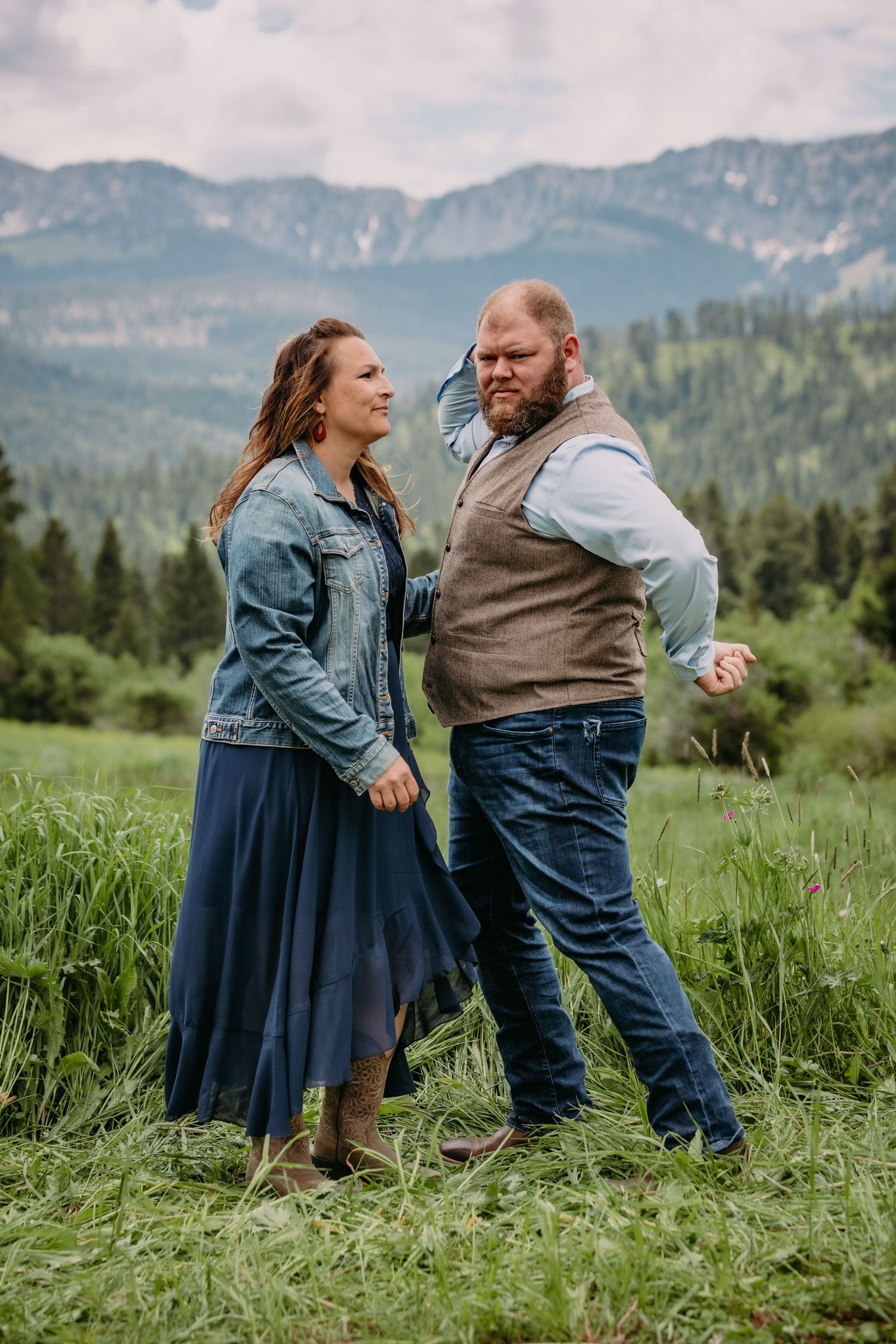 Playful couple dancing in a green field with mountains and forest in the background.