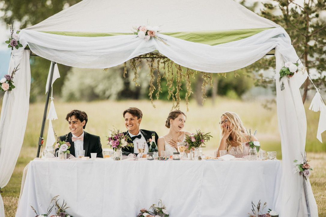 Wedding party seated at head table under white fabric canopy outdoors.