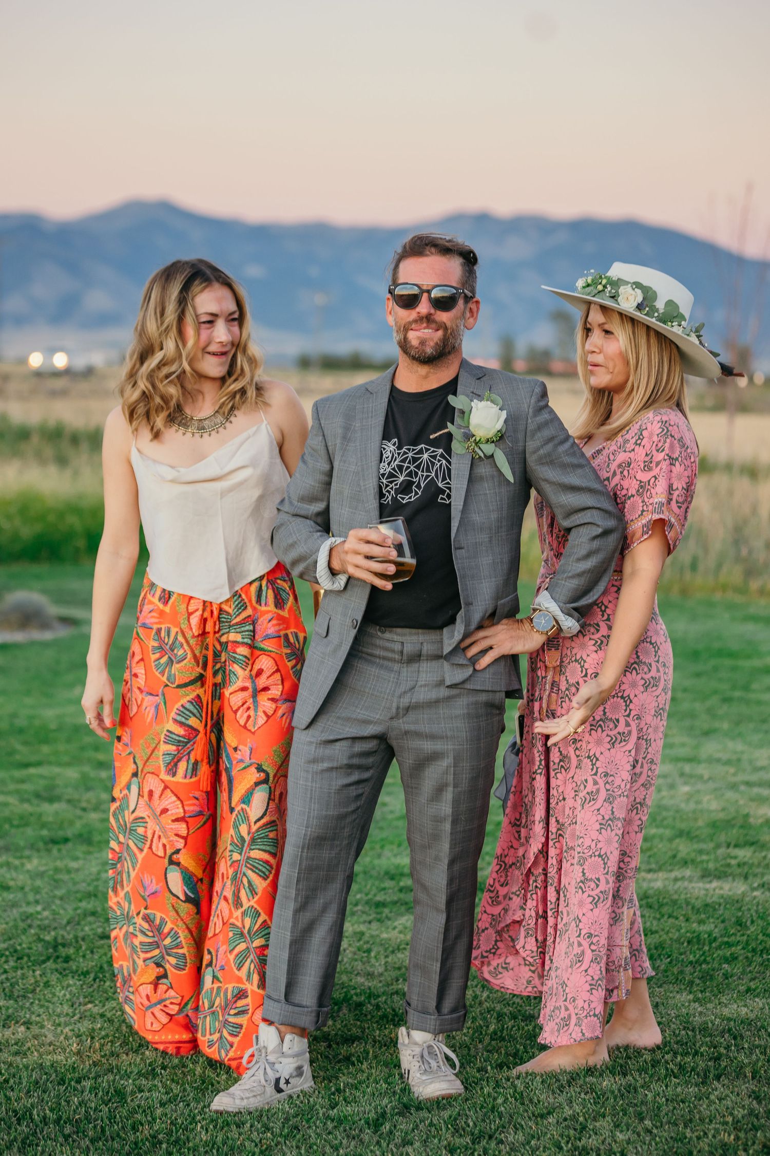 Wedding party poses on grass with colorful floral dresses and casual attire.