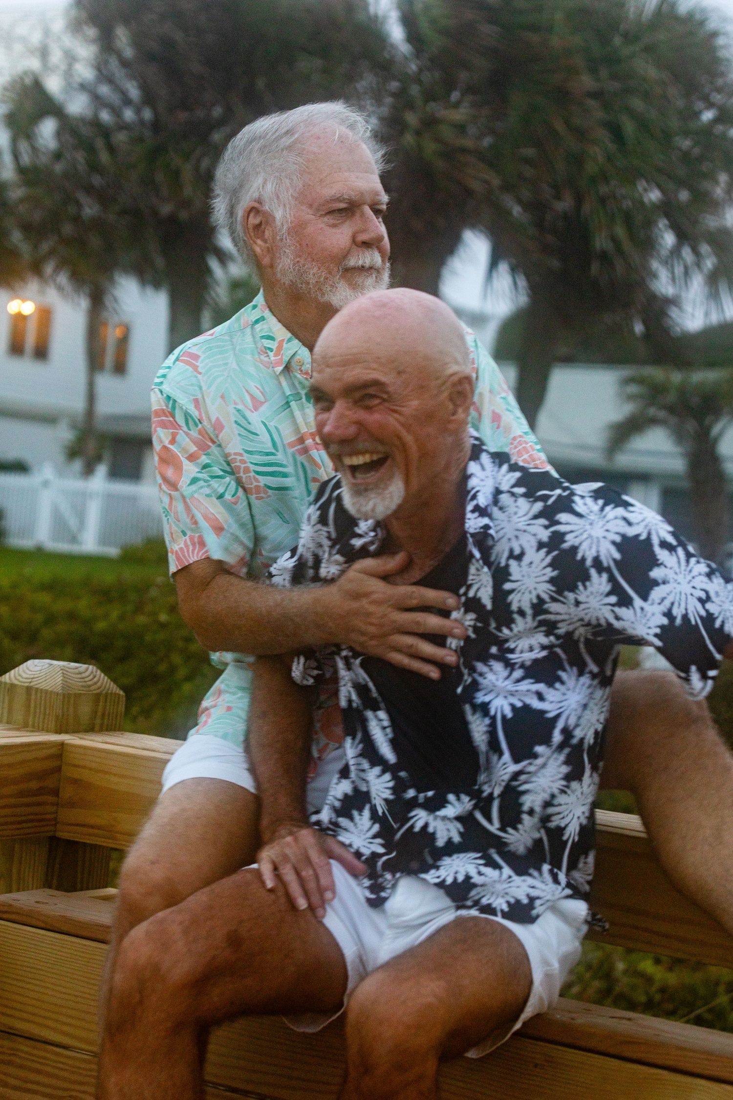 Two older men in tropical shirts laughing together outdoors near palm trees.