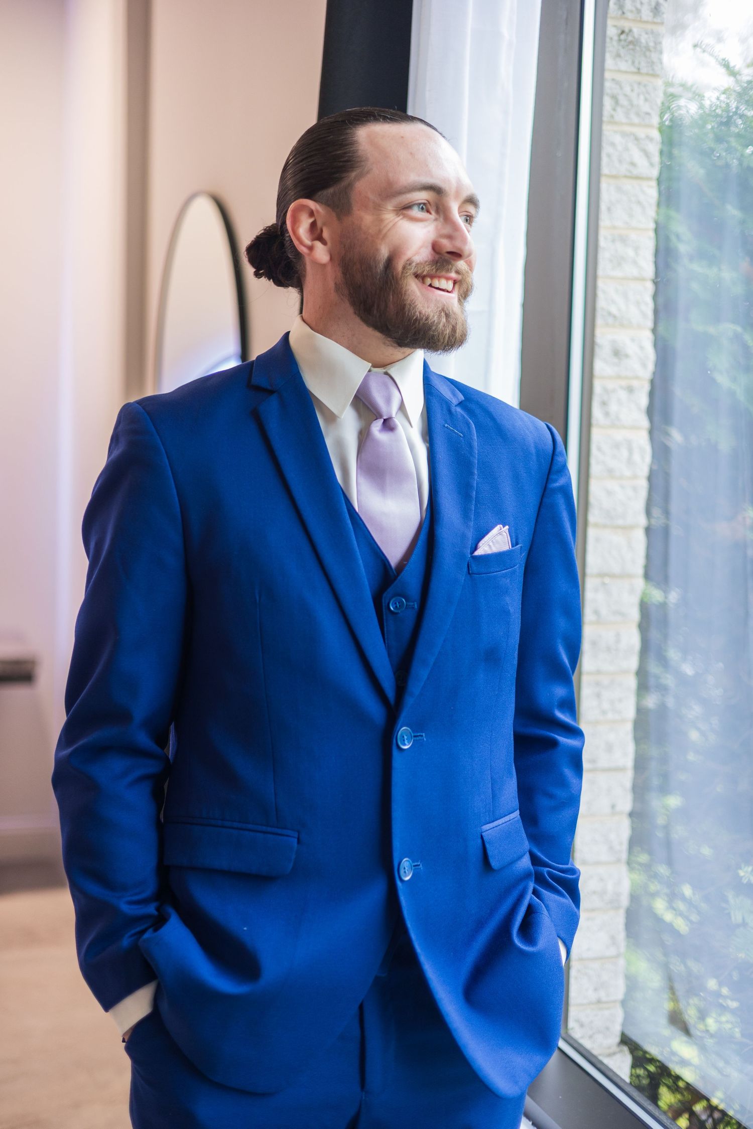 Smiling groom in royal blue three-piece suit with lavender tie stands by window with hands in pockets.