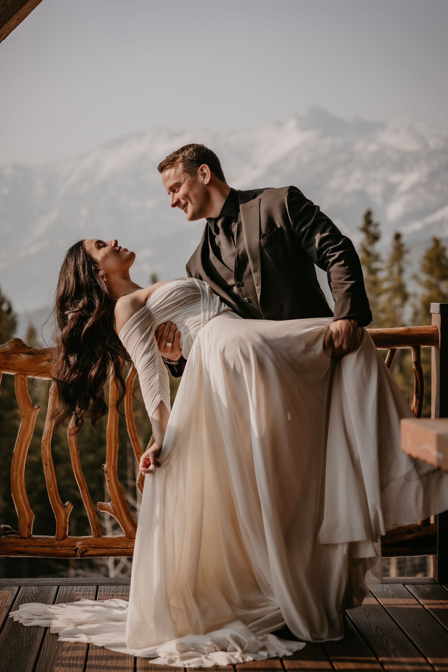 A romantic wedding couple dances on a wooden deck with snowy mountain peaks in the background at sunset.