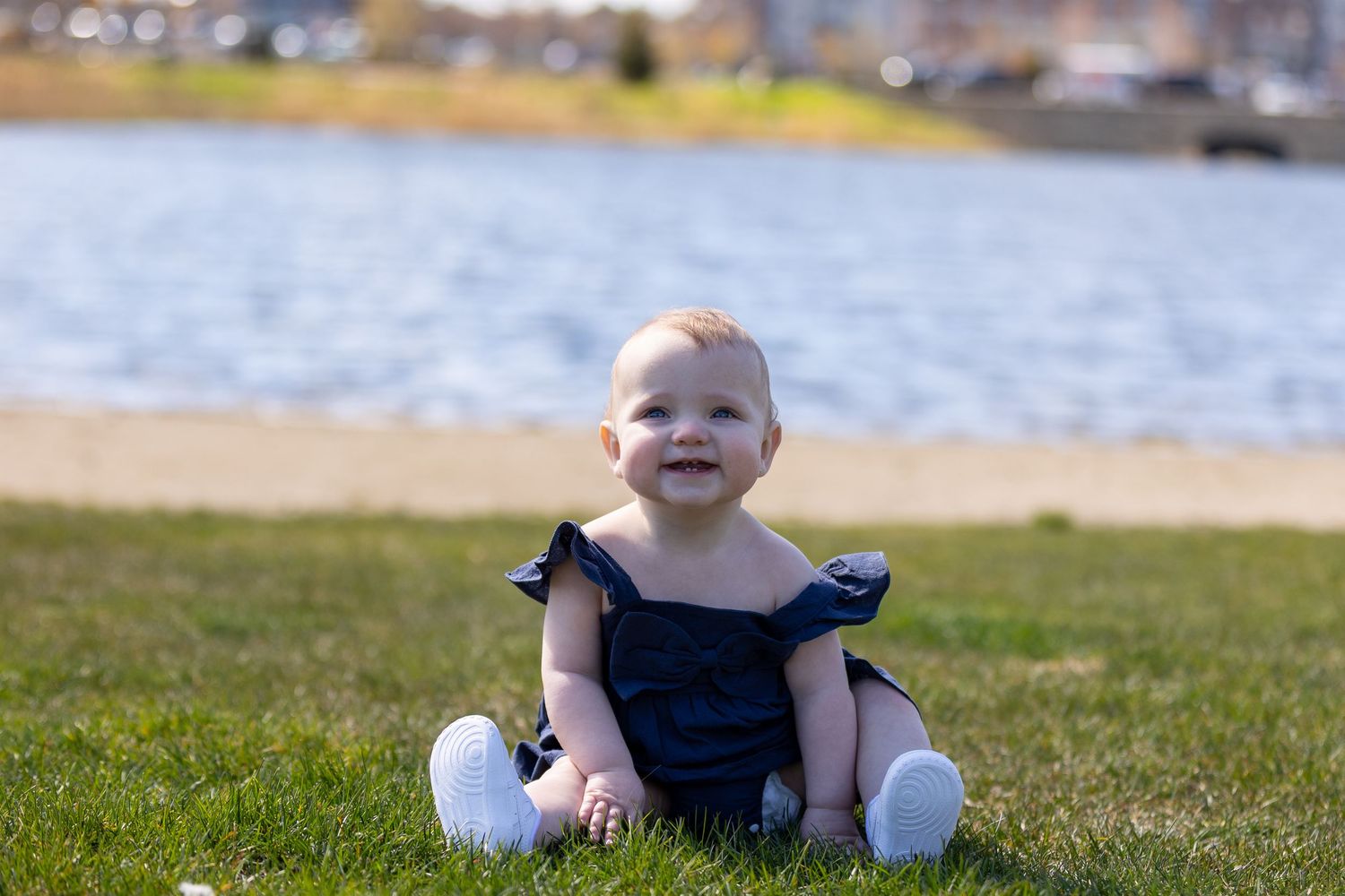 Smiling baby girl in navy blue romper sitting on green grass by a lake on a sunny day.