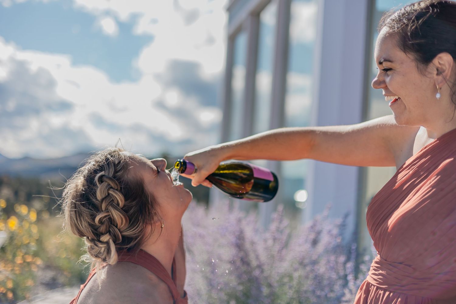 Two women sharing drinks during outdoor wedding preparations with mountain views.