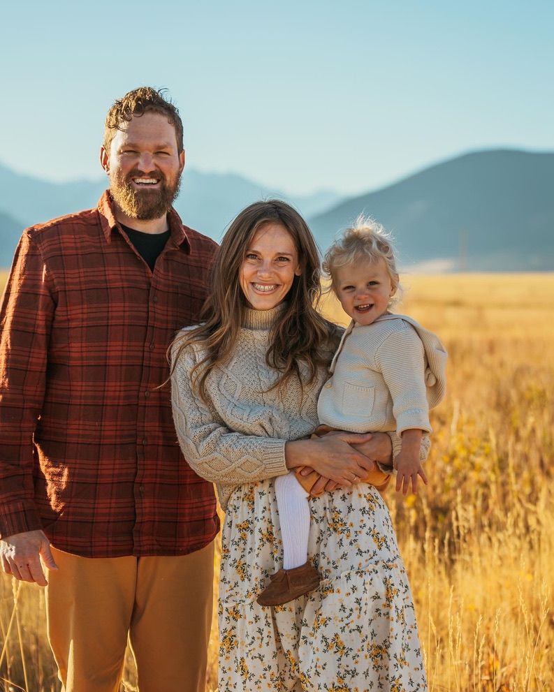 Family stands together in field with mountains and golden grass in background.