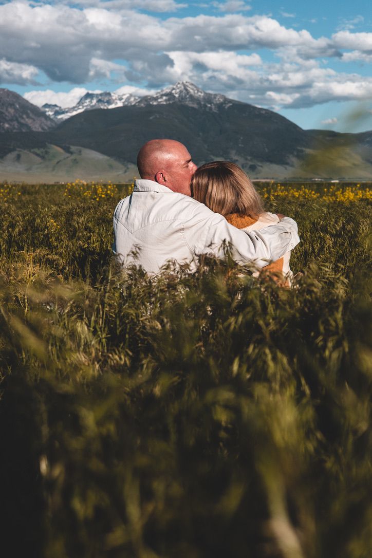 A couple embraces in a field of sunflowers with majestic mountains in the background.