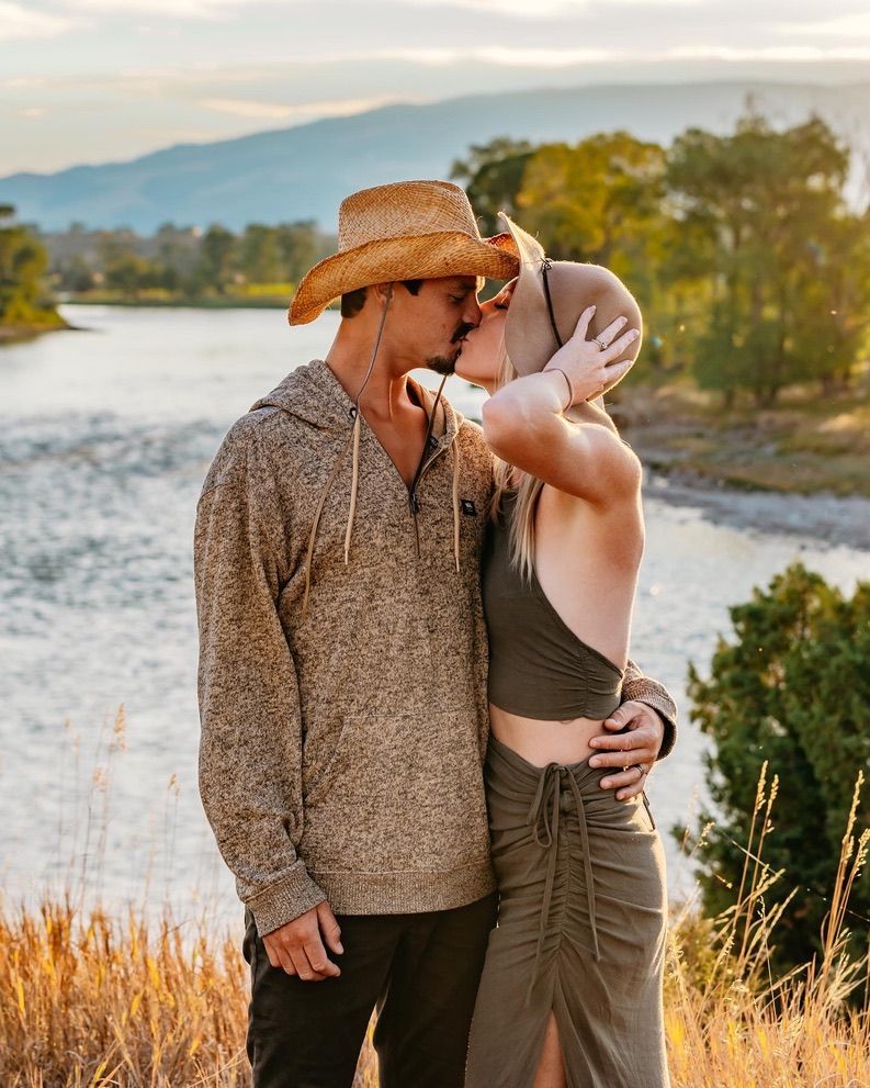 A romantic couple embraces in golden wheat fields with mountains and a lake in the background during sunset.
