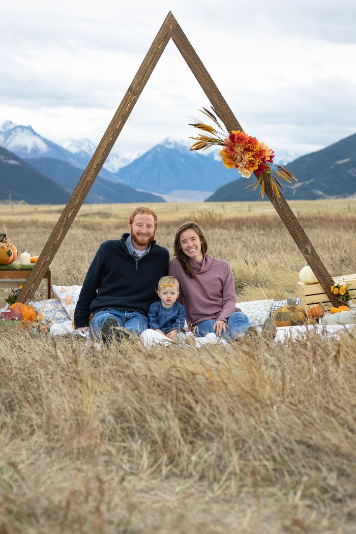 Family picnic setup in a grassy field with wooden triangle frame and mountain backdrop.