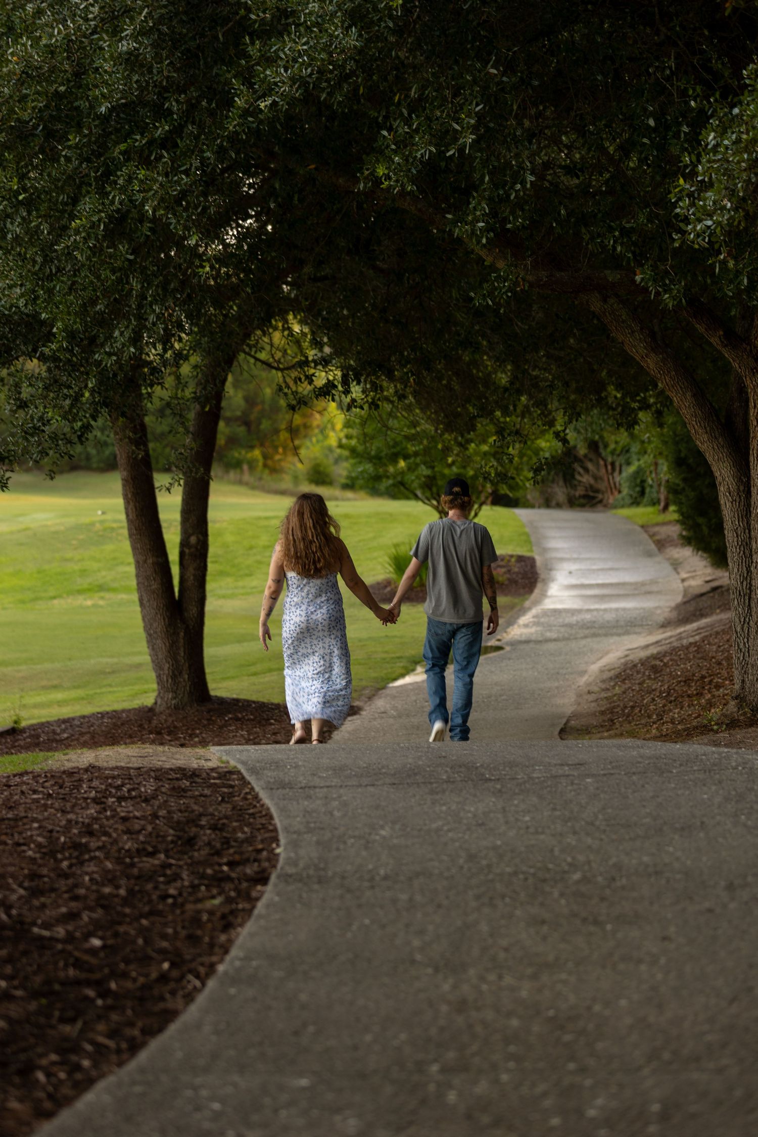 Couple holding hands walking down a winding park path lined with lush green trees on a sunny day.