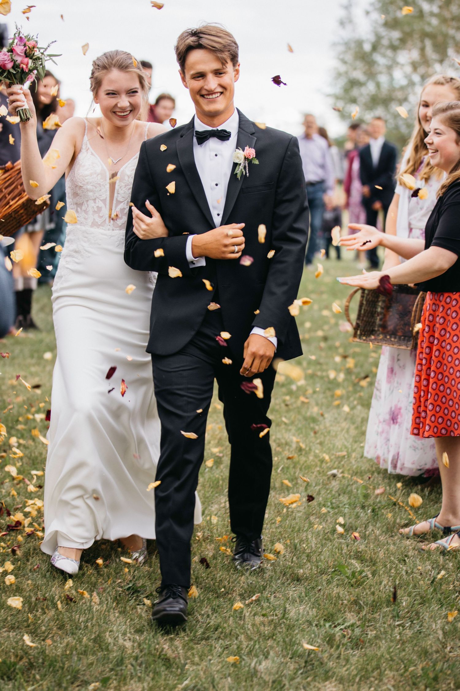 Newlywed couple walks down grassy aisle while guests throw flower petals in joyous outdoor celebration.