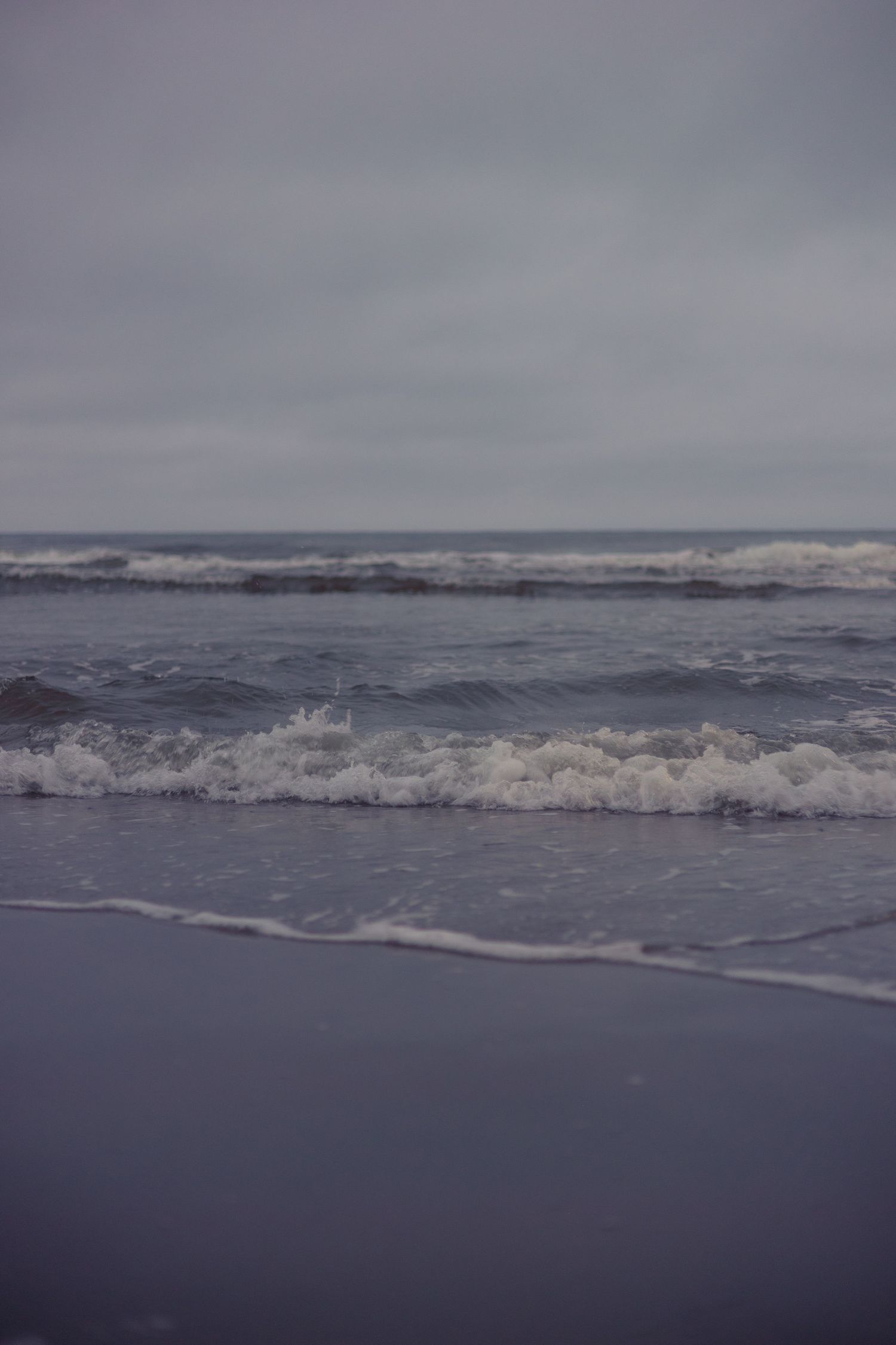 Ocean waves crashing on a dark sandy beach under an overcast sky.
