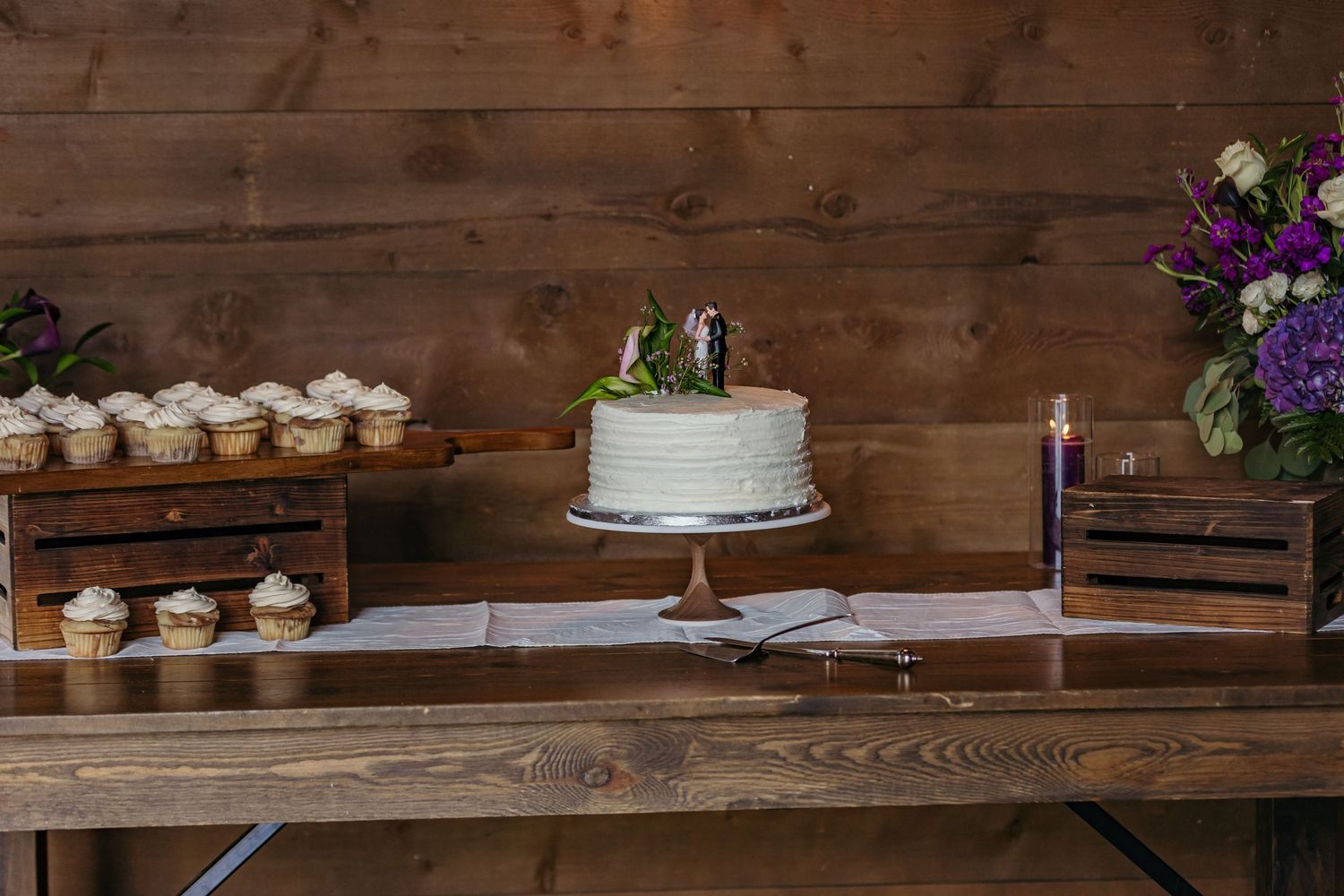 Wedding dessert table with white cake and cupcakes on rustic wooden display.