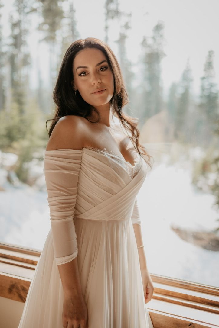 Ethereal winter bridal portrait in flowing off-shoulder cream gown against snowy forest backdrop.