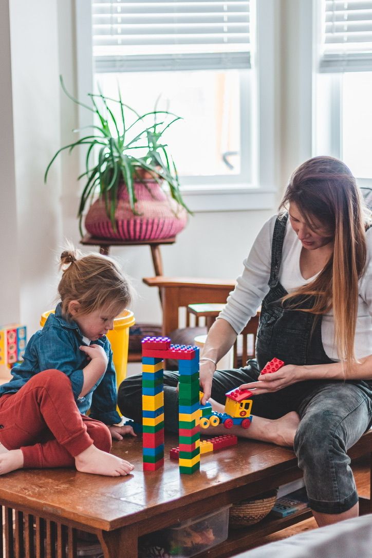 Children and adults play with colorful building blocks on a wooden table near a window with houseplants.
