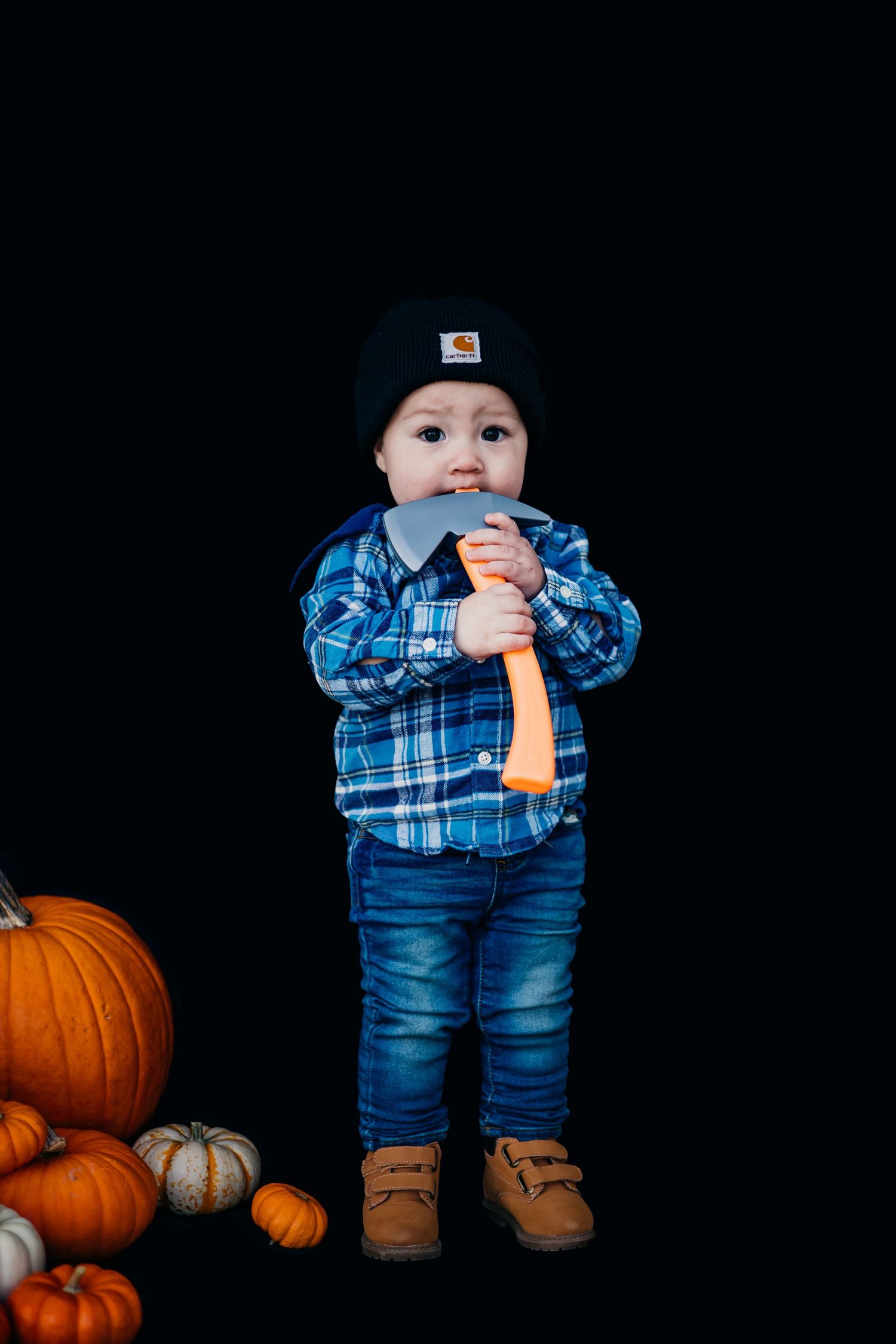 Small child in plaid shirt and beanie stands next to pumpkins holding toy hammer.