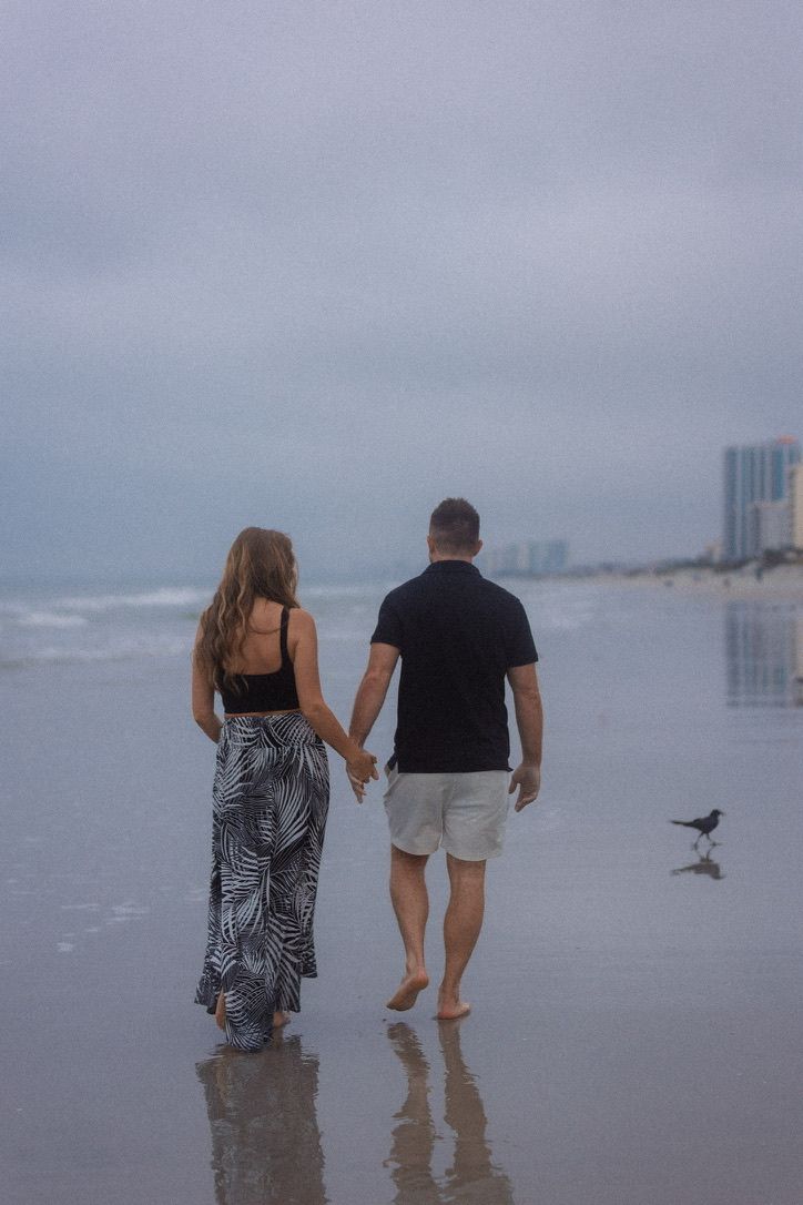 Two people walk along a misty beach with waves and seabirds in the background.