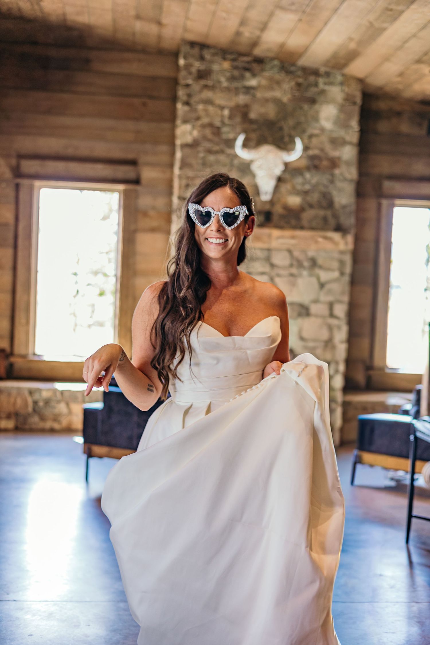 Bride wearing sunglasses dances playfully in rustic venue with stone walls.