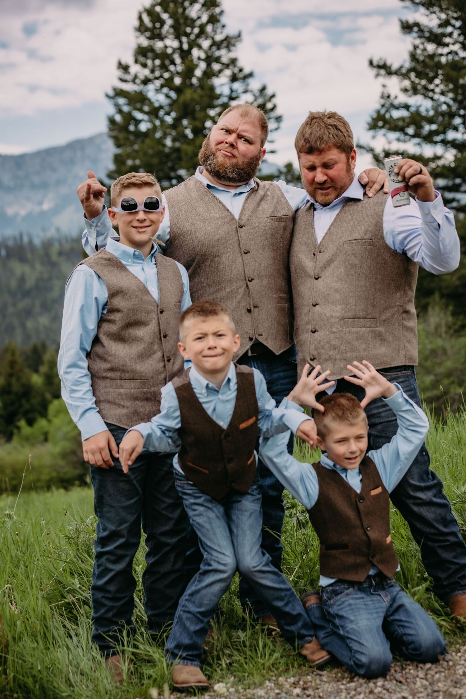 Groomsmen and young boys in matching vests pose playfully against mountain backdrop.