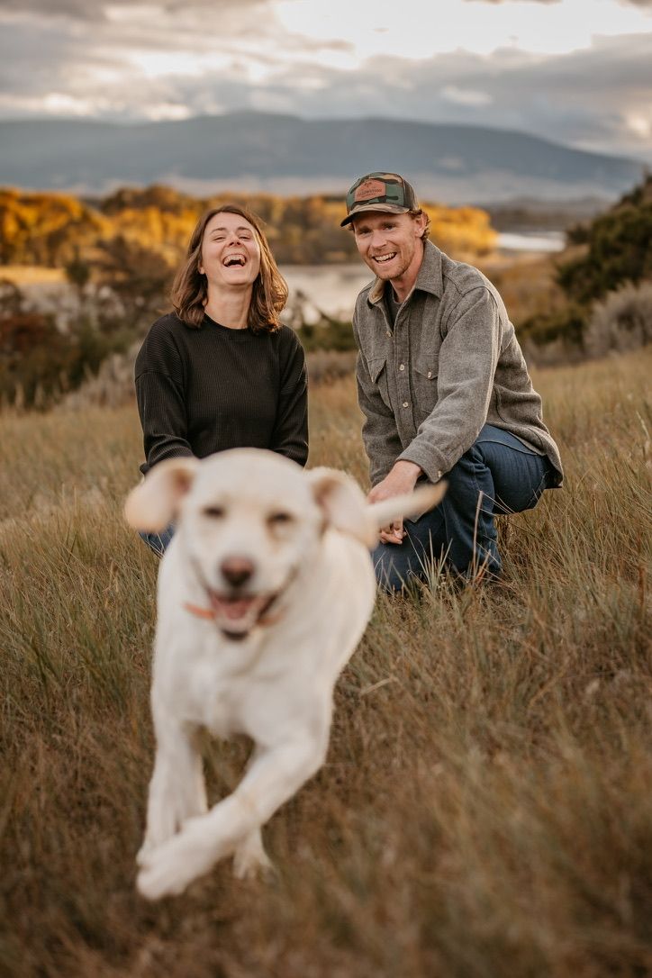 Playful couple laughing with their excited white dog running through field.