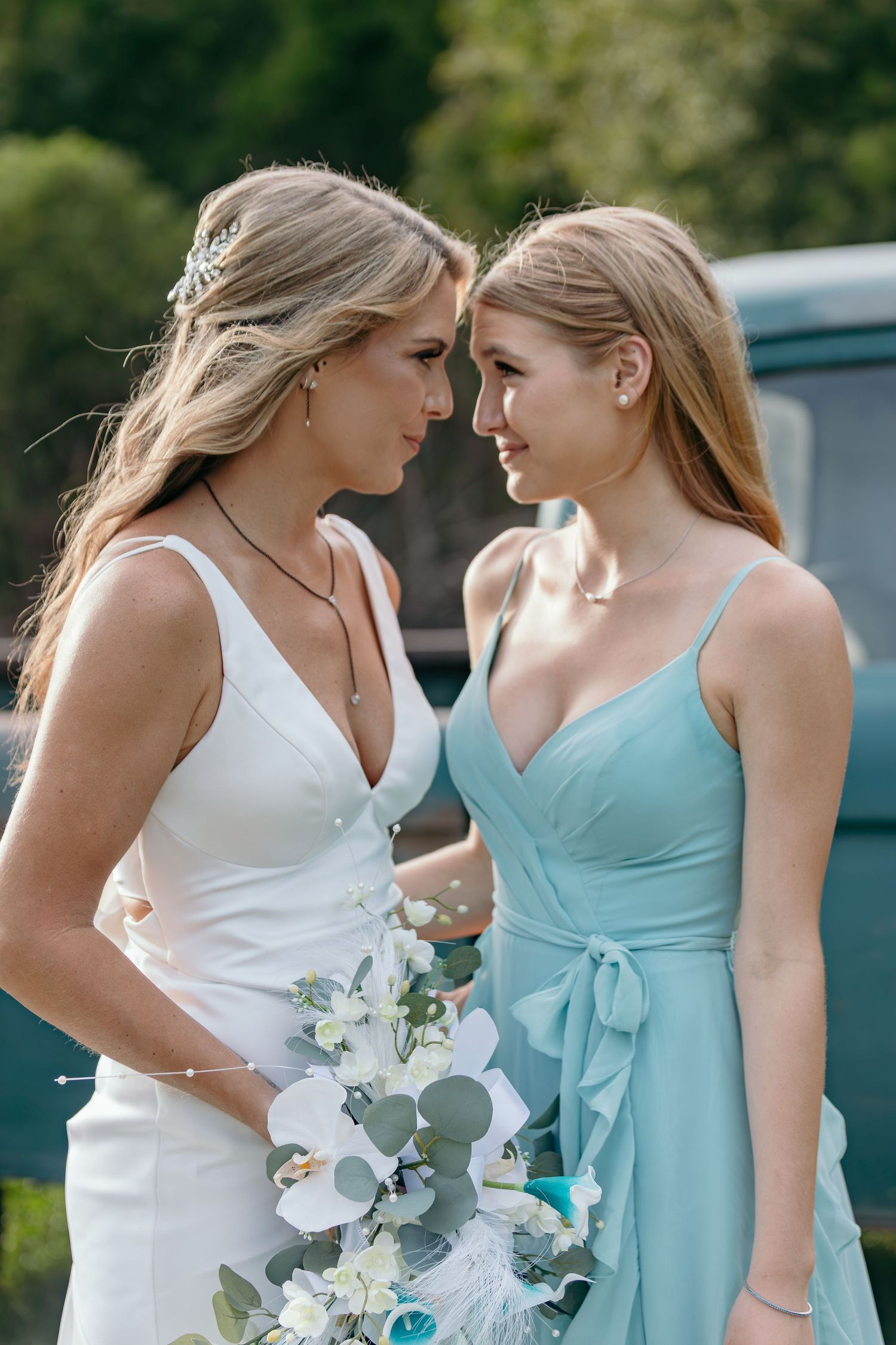 Bride and bridesmaid in white and turquoise dresses pose with orchid bouquet outdoors.