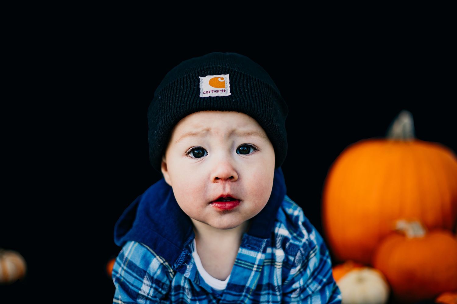Close up of child in blue plaid shirt and black beanie with pumpkins in background.