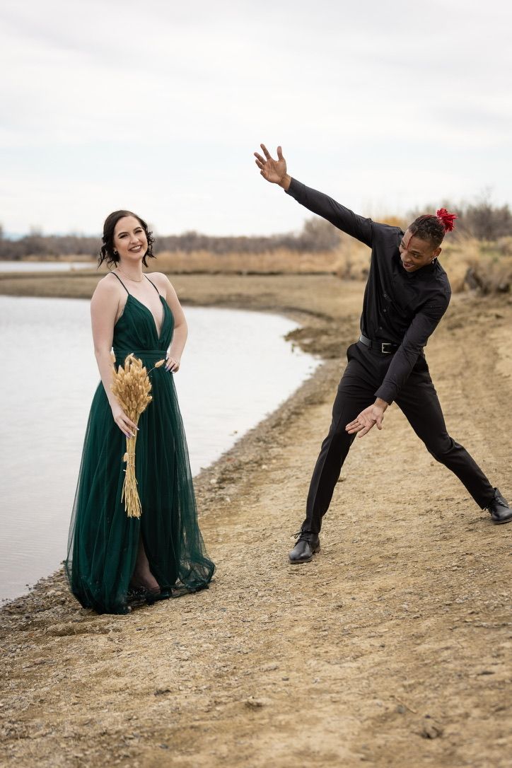 Playful couple in formal attire poses dramatically by riverside.