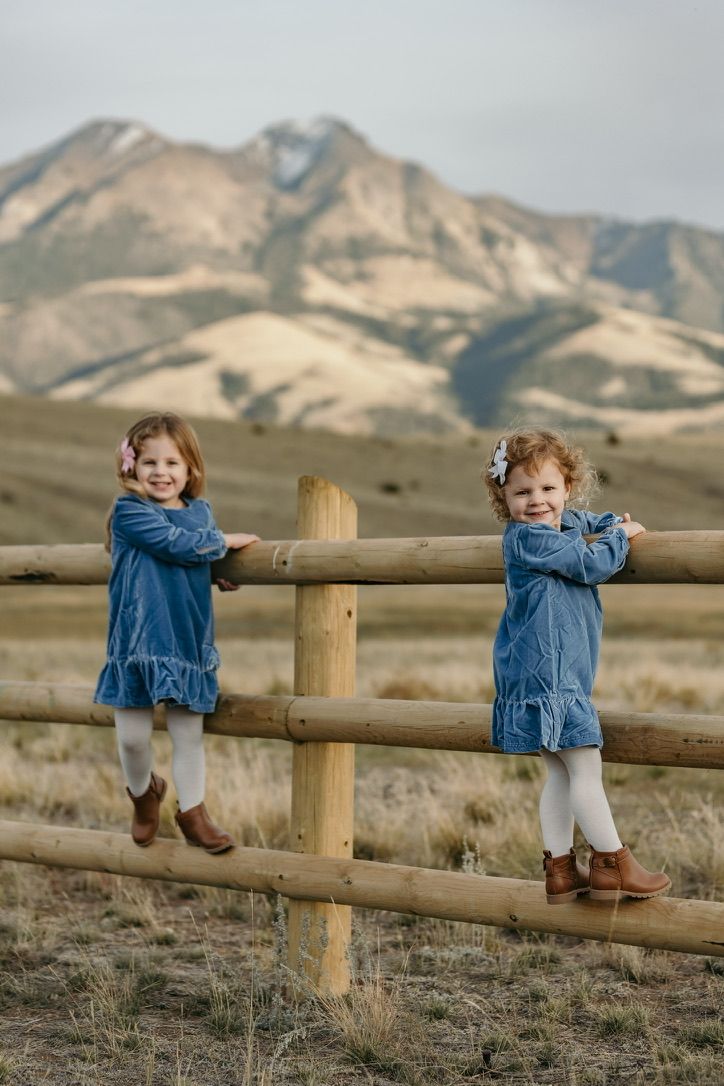 Young child in denim jacket plays on wooden fence with mountains in background.