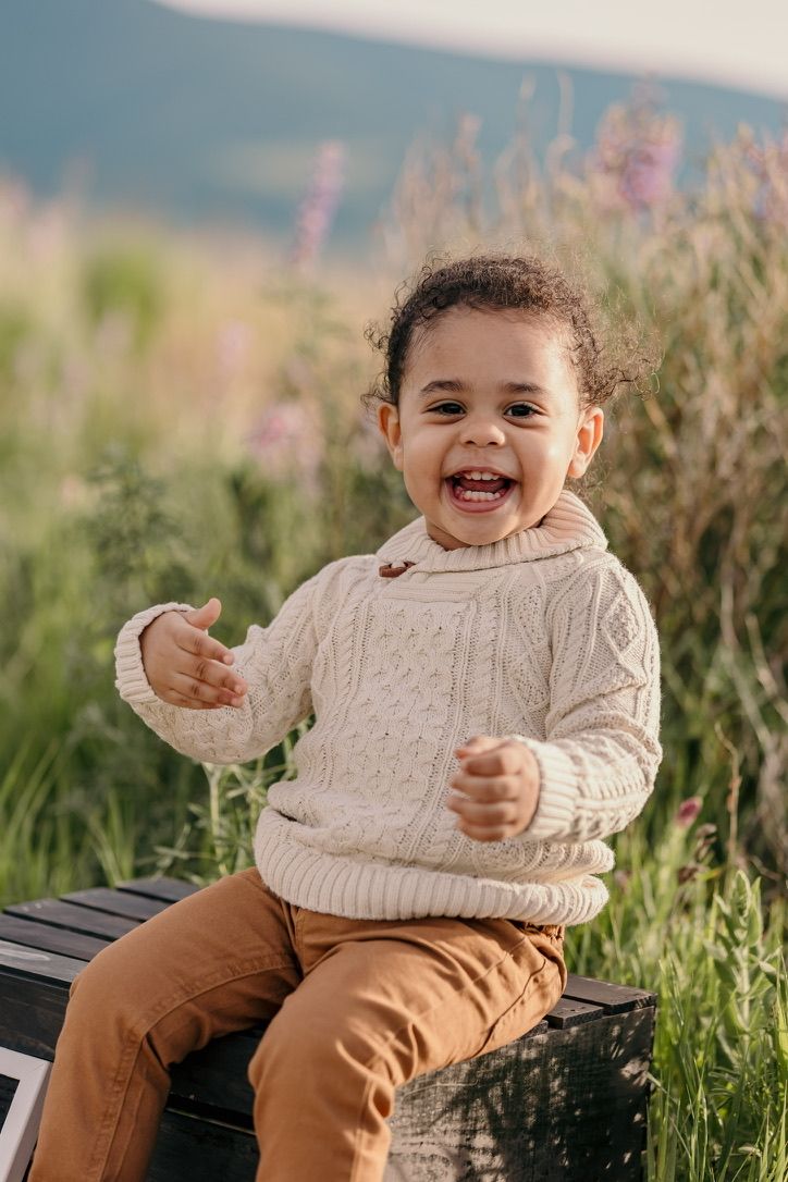 Child wearing cream sweater and brown pants sitting outdoors in grassy field.