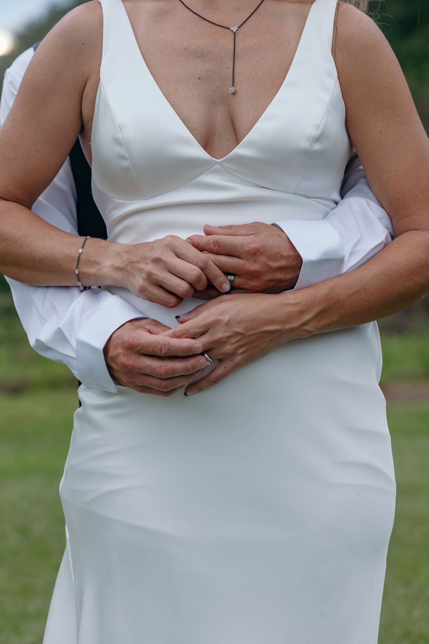 Close-up detail shot of hands and white wedding dress in outdoor setting.