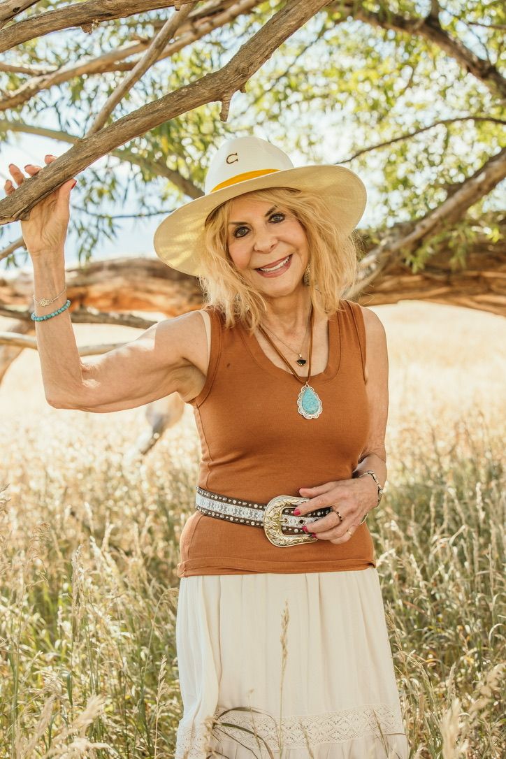 Woman in brown tank top and sun hat poses in nature with a turquoise necklace against a sunlit background.