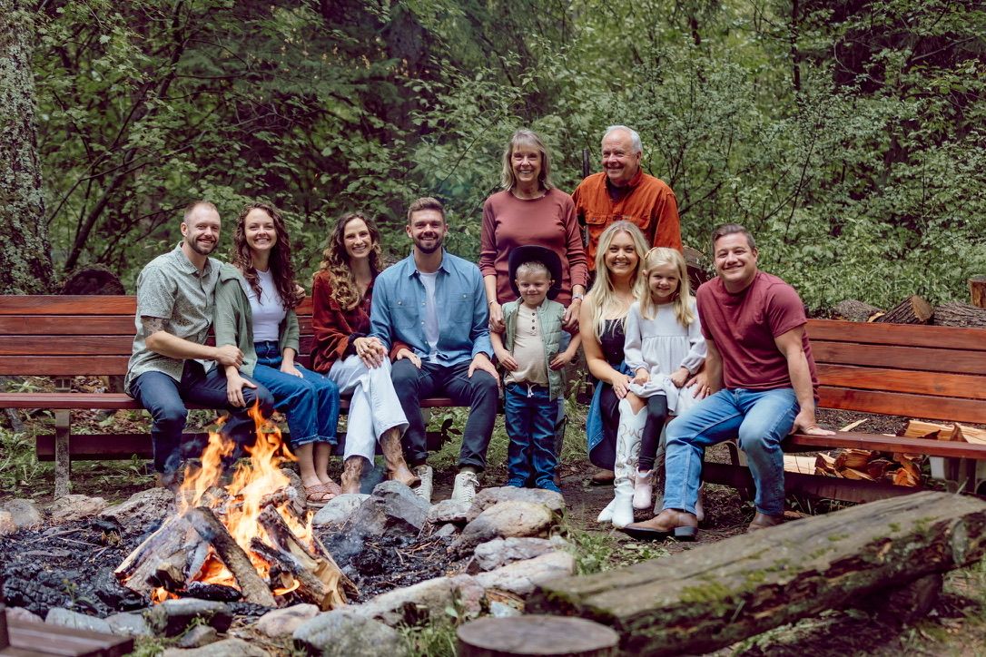 Large group gathered around outdoor campfire on wooden benches in wooded setting.