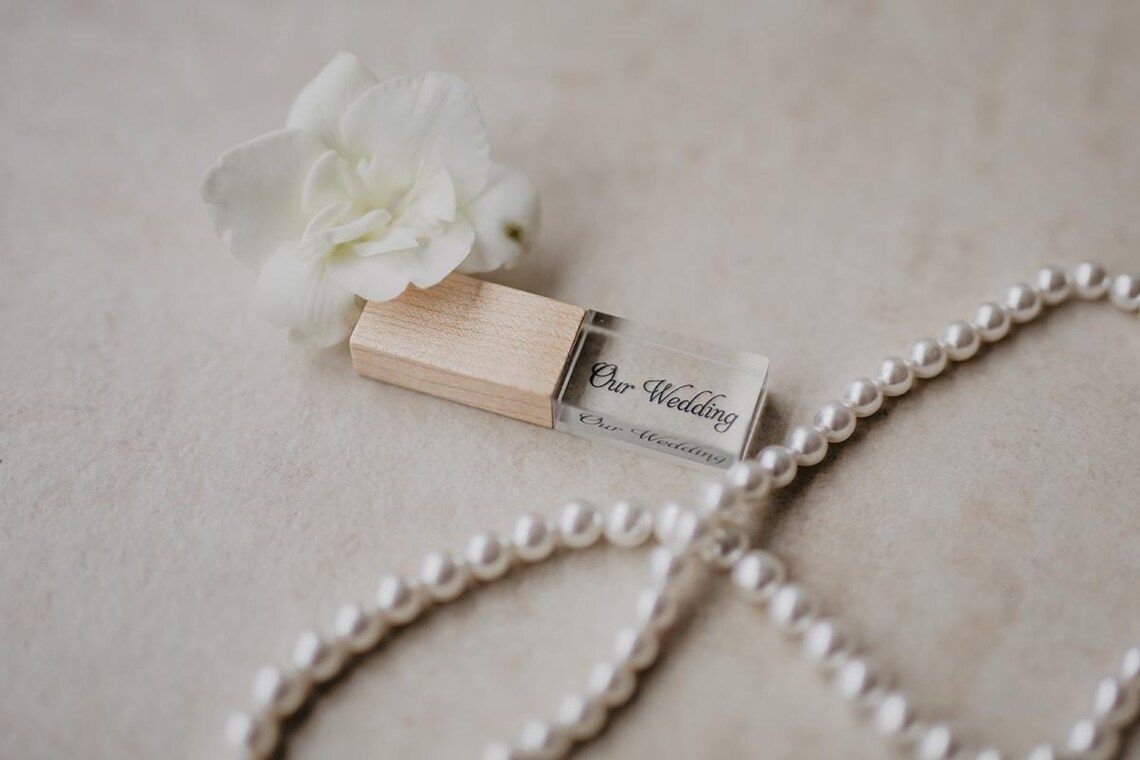 A wooden USB flash drive rests elegantly beside a white flower and pearl necklace on a beige surface.