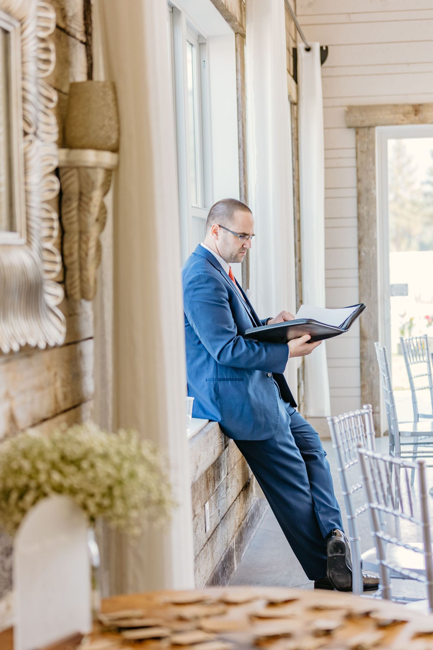 Person in blue suit reads from a book on a white porch with silver chairs.