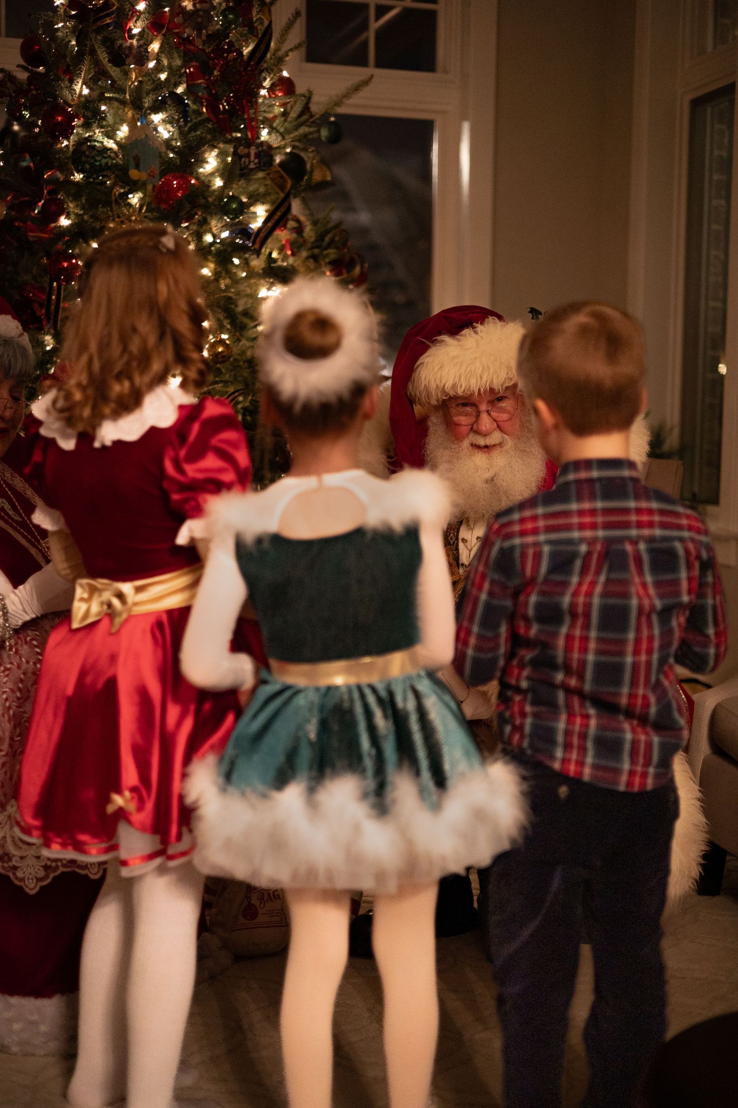 Children in festive costumes meet Santa Claus beside a decorated Christmas tree in a cozy holiday setting.